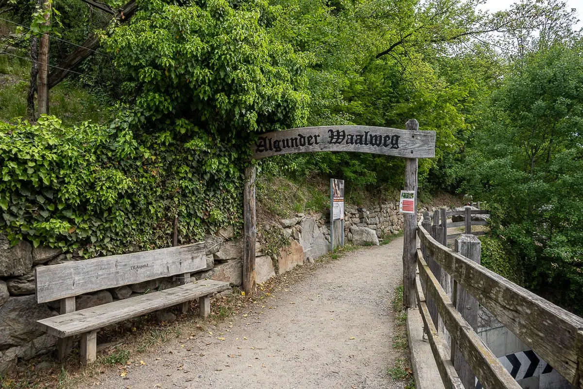 Ein Feldweg führt durch einen hölzernen Torbogen mit der Aufschrift „Algunder Waalweg“. Der Weg ist von Holzgeländern gesäumt, links davon stehen üppig grüne Bäume, Steinmauern und eine Bank. Informationsschilder heißen die Besucher auf diesem malerischen Wanderweg, dem Algunder Waalweg, willkommen.