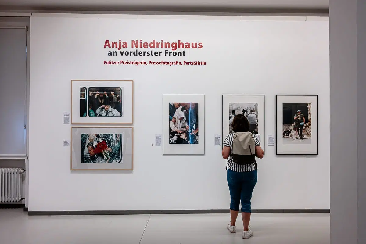 Eine Frau steht vor gerahmten Kriegsfotografien von Anja Niedringhaus in einer Galerie im Rahmen der Anja-Niedringhaus-Ausstellung in Oberhausen. Der Wandtext lautet „Anja Niedringhaus an vorderster Front“ und verweist auf die Ausstellung; neben jedem Foto befinden sich mehrere beschreibende Beschriftungen.