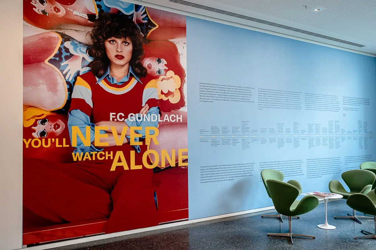 Ein großes, farbenfrohes Wandbild zeigt eine Frau in einer roten Latzhose, hinter der Clownfiguren zu sehen sind. In kräftigem Gelb steht dort: „YOU’LL NEVER WATCH ALONE F.C. GUNDLACH.“ Daneben sind auf den blauen Wänden Textabschnitte zu lesen. Im Vordergrund stehen drei grüne Stühle und ein runder Tisch.