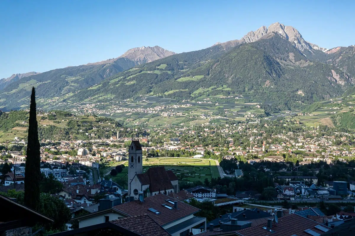 Das malerisches Städtchen Marling mit einer Kirche und Gebäuden mit roten Dächern liegt in einem grünen Tal, umgeben von sanften Hügeln und hohen, bewaldeten Bergen unter einem strahlend blauen Himmel. Die Landschaft ist üppig und weitläufig und spiegelt die Schönheit einer sonnigen, ländlichen Region wider.