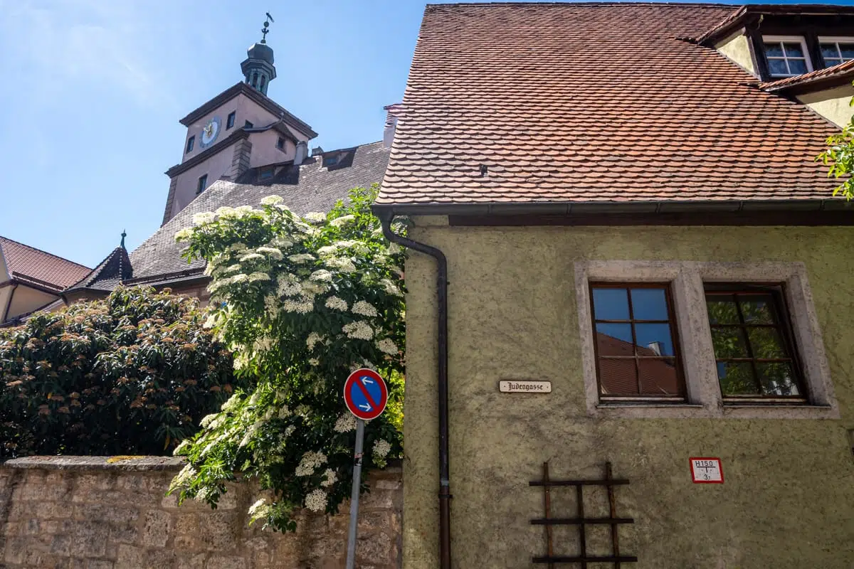 Ein hellgrünes Haus mit rotem Ziegeldach steht neben einem blühenden Strauch und einer Steinmauer. Es ist ein Schild mit dem Wort "Judengasse" angebracht. Dahinter erhebt sich ein Kirchturm mit Uhr und grüner Turmspitze. Davor steht ein blau-rotes Parkverbotsschild, und über dem Ganzen erstreckt sich ein strahlend blauer Himmel.