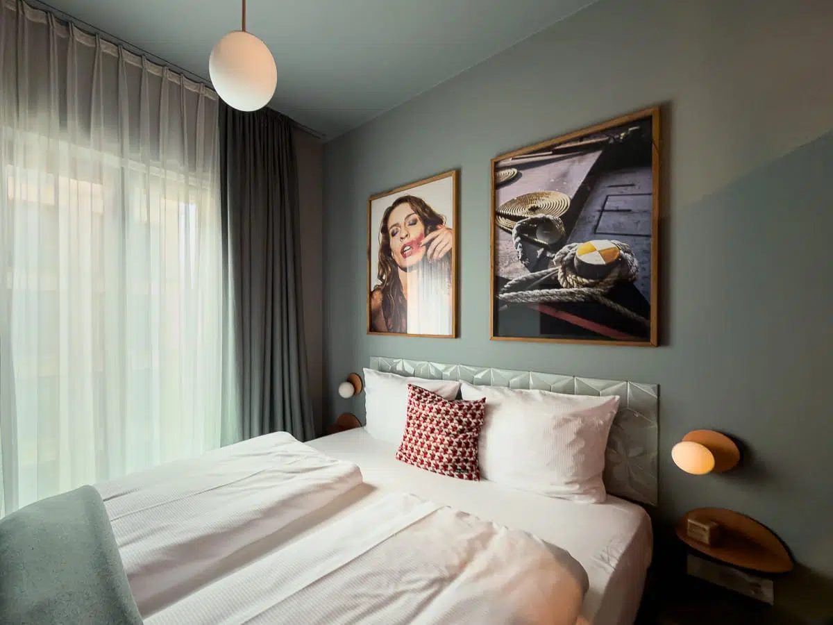 An einer grauen Wand steht ein ordentlich gemachtes Bett mit weißer Bettwäsche, darüber hängen zwei gerahmte Fotos. Das eine Foto zeigt eine Frau, das andere maritime Motive. Ein geometrisches Kissen sorgt für einen Farbtupfer. Hohe Fenster sind mit Vorhängen verhängt, und zu beiden Seiten des Bettes befinden sich moderne Wandleuchten. Über dem Bett hängt eine runde Deckenleuchte, die den gemütlichen Raum abrundet, ein Hotelzimmer im „lyf East Frankfurt“.